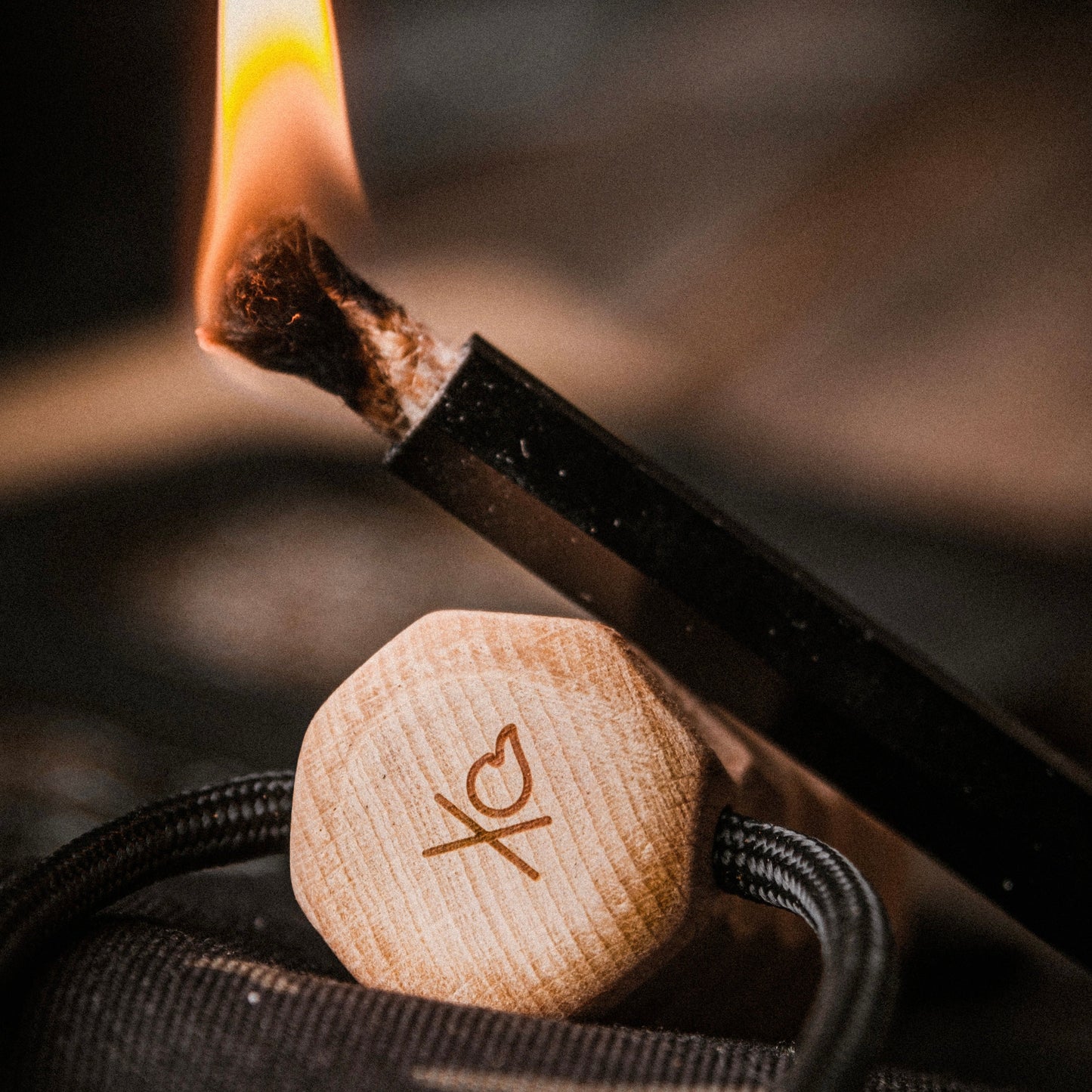 A close-up of an incense stick being lit with the Überleben Kräftig Grip (PRO)—The Heavy-Duty Ferro Beast by Überleben, while a wooden cap engraved with XO and a heart symbol sits in focus. A blurred background completes the scene.
