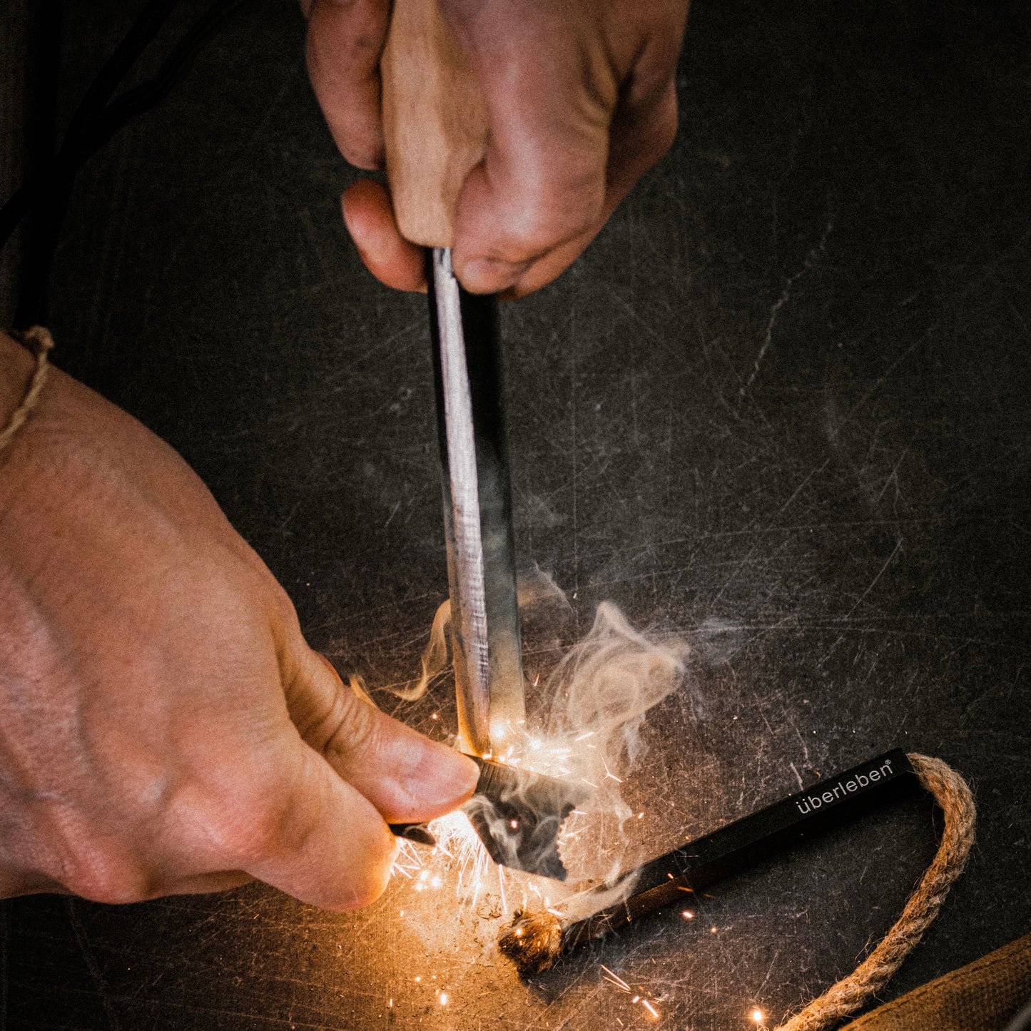 Two hands strike the Überleben Kräftig Grip (PRO) – The Heavy‑Duty Ferro Beast with a knife, creating sparks and igniting tinder on a dark surface, showcasing this reliable Überleben fire starter in action.