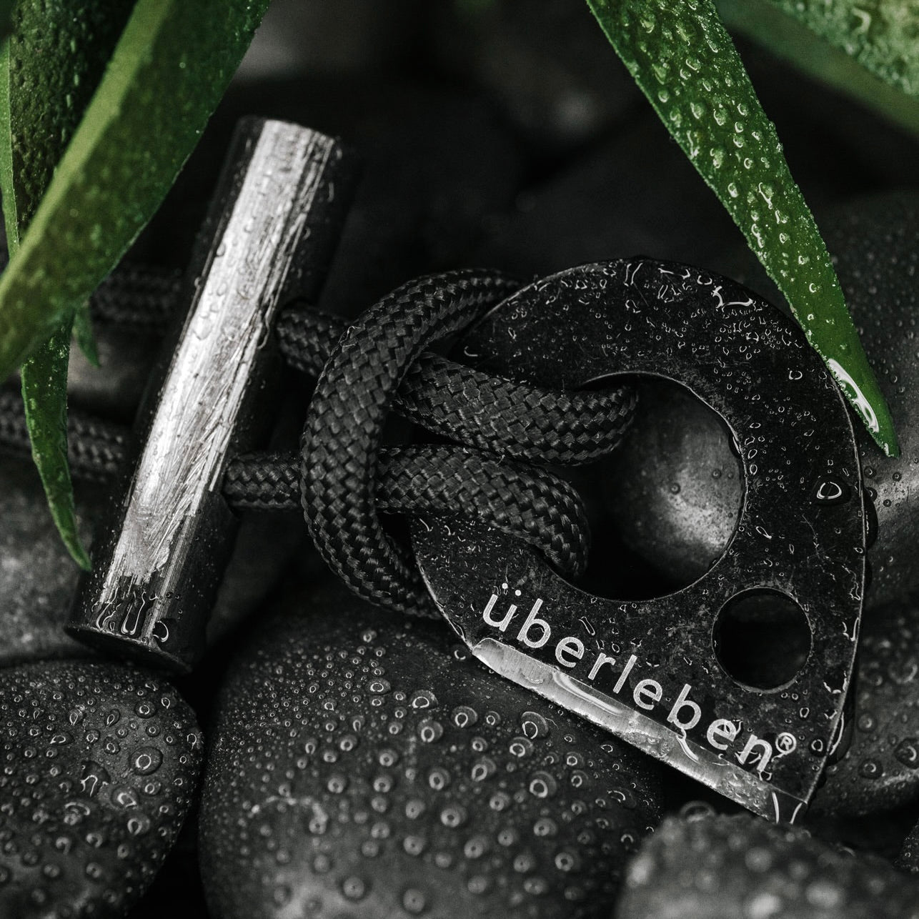 A close-up of the Überleben Leicht – Wearable Fire Starter by Überleben, a black metal tool with black paracord, resting on wet stones and surrounded by green leaves with water droplets.