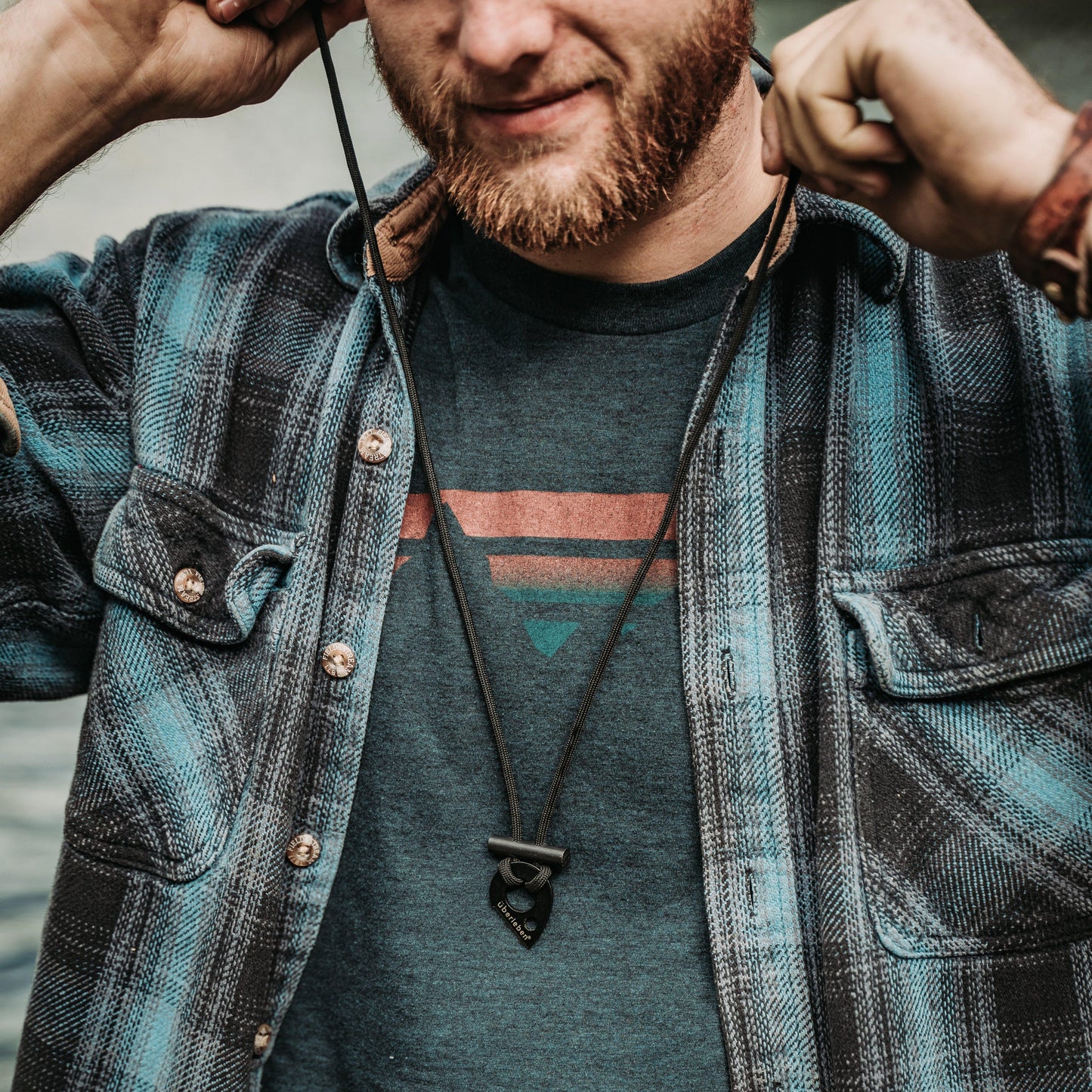 A man with a reddish beard and blue plaid shirt adjusts his Überleben Leicht – Wearable Fire Starter pendant by Überleben. The focus is on his torso and hands.