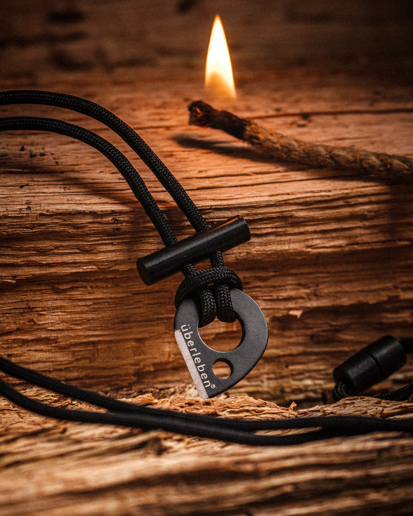 An Überleben Leicht – Wearable Fire Starter with überleben engraved lies on rough wood, as a lit stick flickers in the background.