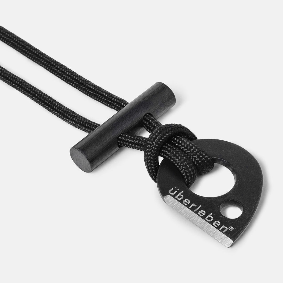 Close-up of the Überleben Leicht – Wearable Fire Starter by Überleben, a black tool with a metal striker engraved überleben, attached to a black cord and featuring a cylindrical micro ferro rod for creating sparks.