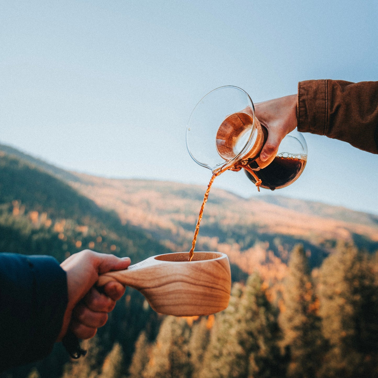 Someone pours coffee from a glass carafe into the Überleben The Lore Oversized Hardwood Kuksa held by another, with forested mountains and a clear sky in the background.