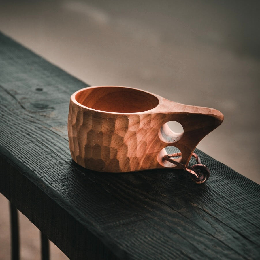 The Überleben Original Kuksa – The Classic Nordic Cup by Überleben is a hand-carved hardwood cup with a handle and leather strap, shown on a dark wooden railing in soft natural light—a beautiful tribute to Nordic tradition.
