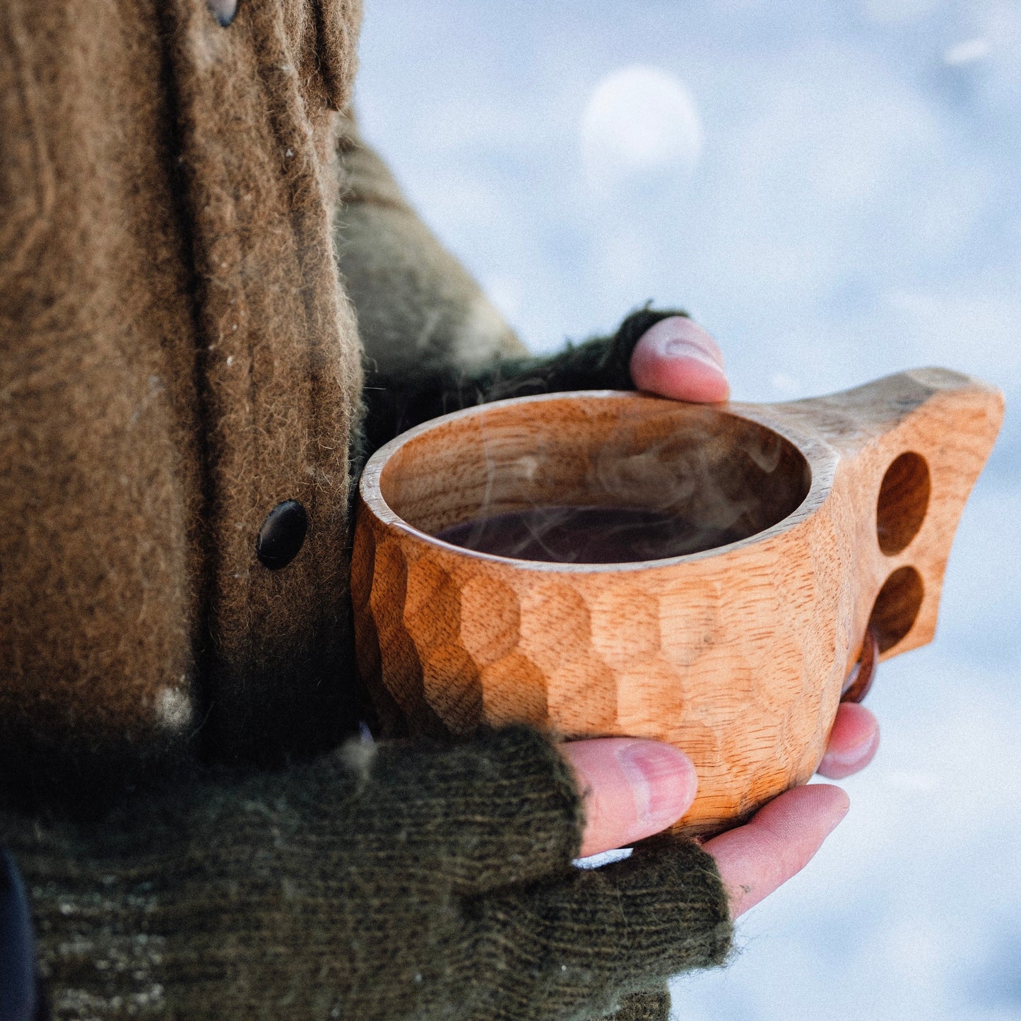 Wearing a brown coat and green knit gloves, someone holds the Überleben Original Kuksa – The Classic Nordic Cup by Überleben, a hand-carved hardwood mug with a textured finish, against snowy scenery, capturing true Nordic tradition.