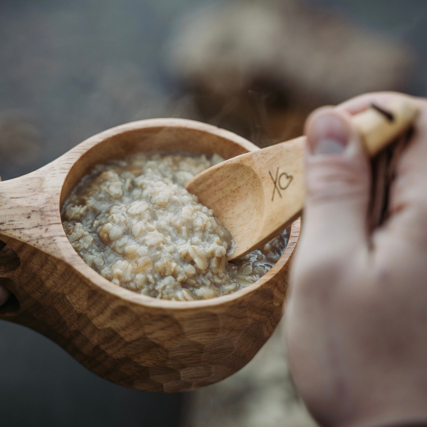 A hand grips the Überleben Original Kuksa – The Classic Nordic Cup, filled with steaming oatmeal, as another hand scoops some up with a wooden spoon.