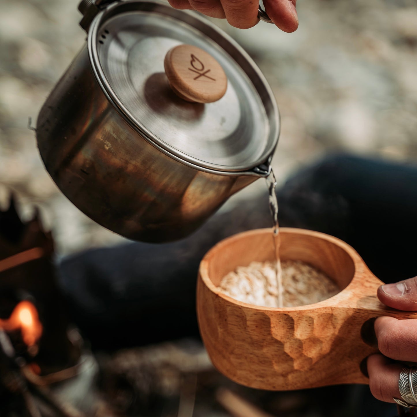 A close-up of someone pouring hot water from a metal kettle into the Überleben Original Kuksa – The Classic Nordic Cup by Überleben, filled with oats, with a campfire glowing in the background—honoring Nordic tradition.