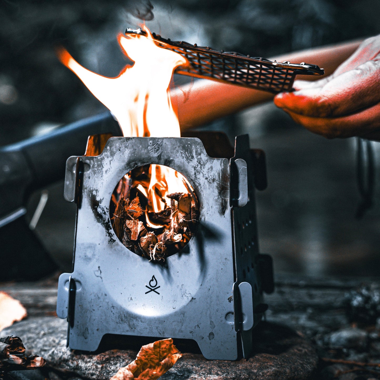 The Überleben Stöker Flatpack Stove by Überleben is a portable wood-burning stove that shows visible flames fueled by leaves, with a hand holding a grill grate above, set against an outdoor backdrop—an essential survival tool.