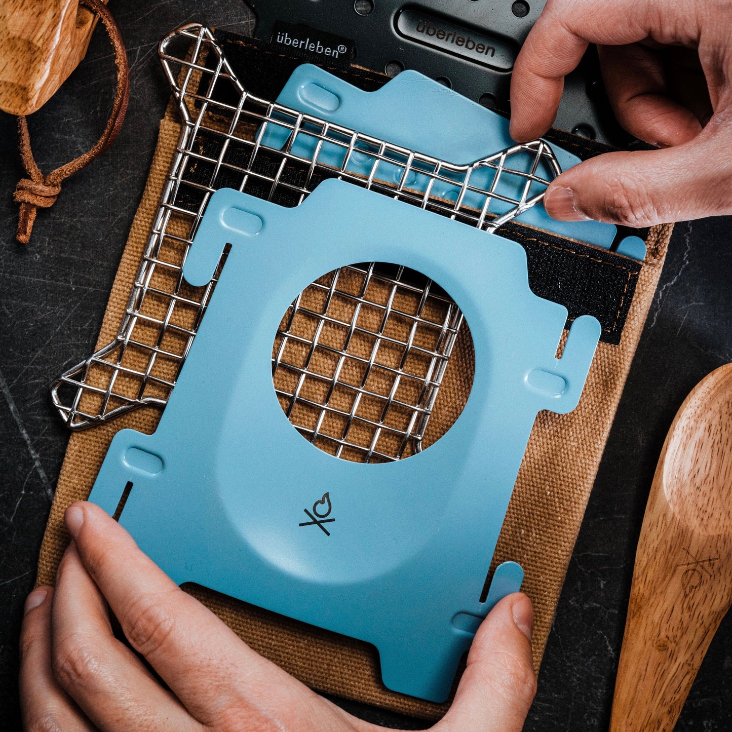 Two hands assemble the Überleben Stöker Flatpack Stove by Überleben—a portable blue camp stove with a metal grid, resting on a brown pouch beside a wooden spoon and lighter. Perfect for outdoor adventures and survival situations.