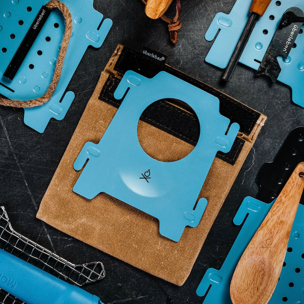 Flat lay of Überleben Stöker Flatpack Stove blue grilling tool accessories, a brown canvas pouch with Velcro flap, and wooden-handled utensils by Überleben on a dark surface. Central blue tool features a circular cutout and minimalist logo.