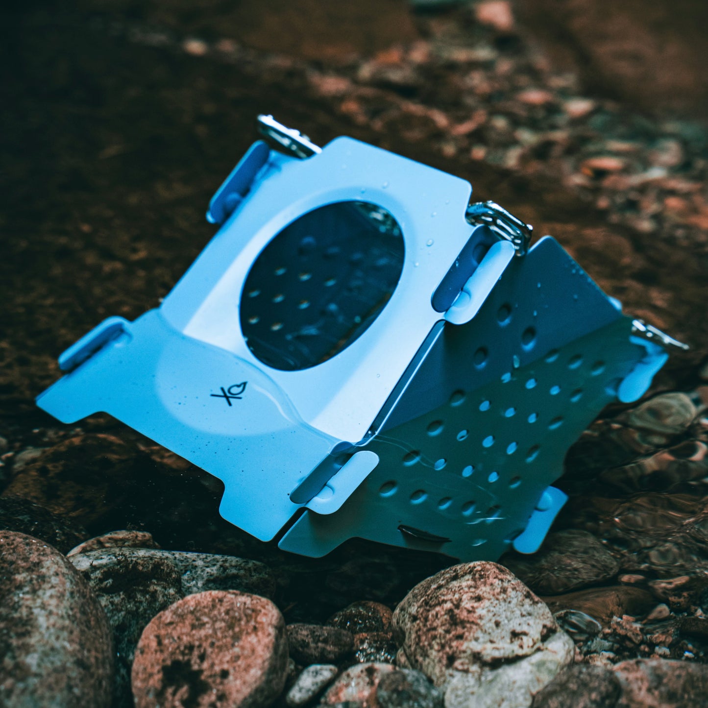 The Überleben Stöker Flatpack Stove by Überleben, a blue foldable wood-burning stove with metal hinges and a circular opening, sits on wet, rocky ground outdoors.