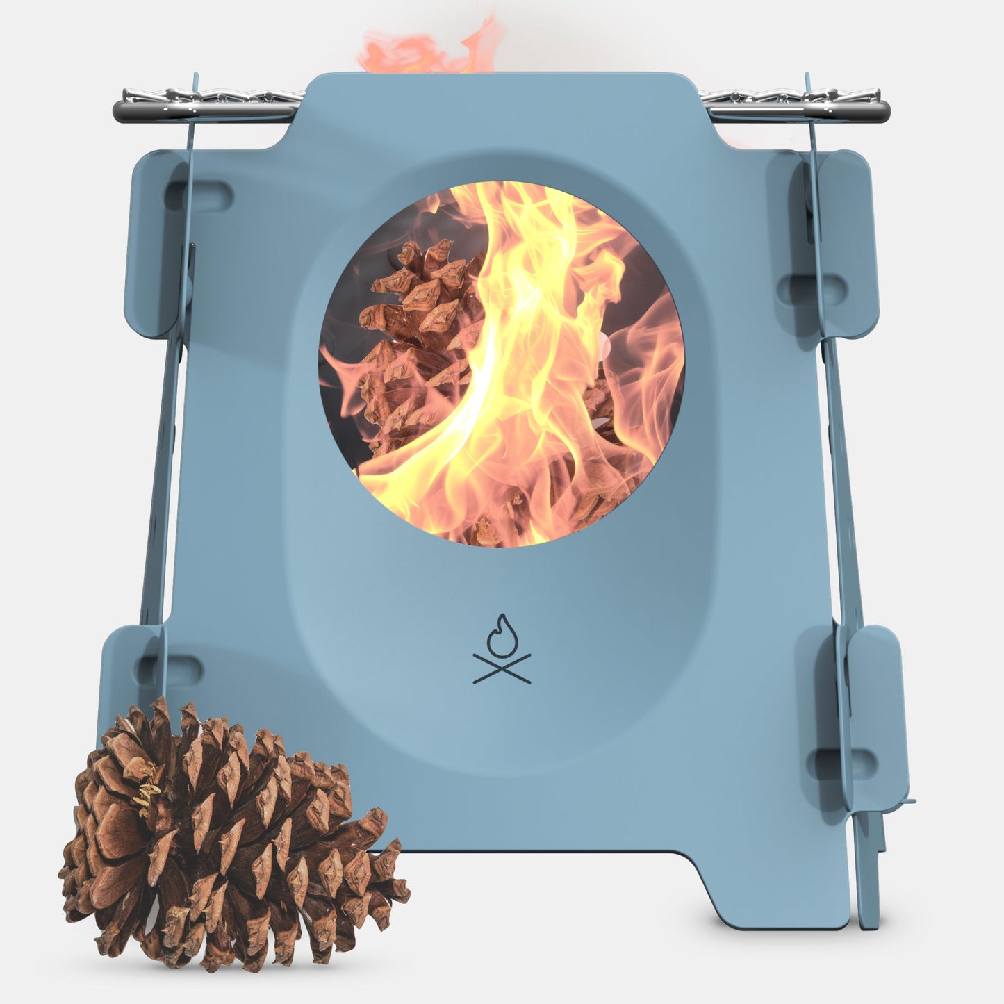 The Überleben Stöker Flatpack Stove by Überleben is a compact blue camp stove with a circular window displaying flames burning pinecones. A large pinecone sits in front of this portable survival tool, photographed on a white background.