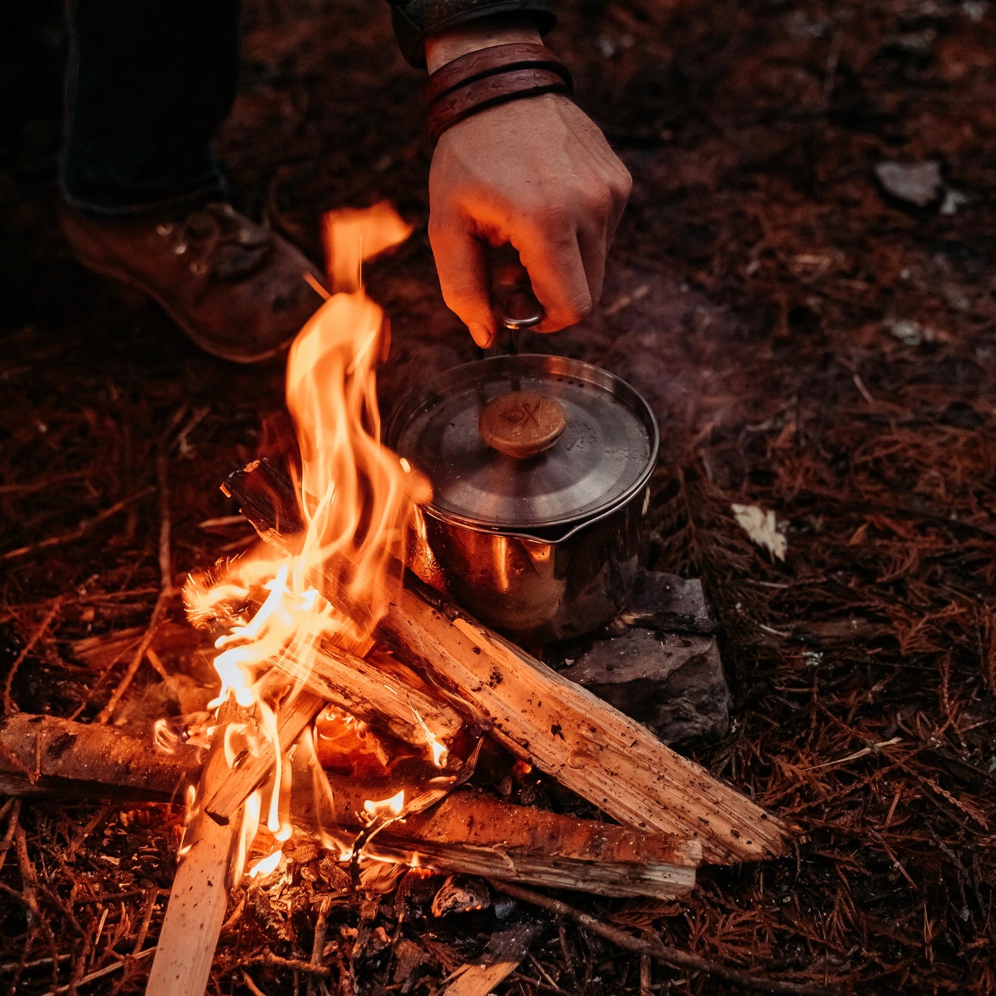 A hand lifts the lid off an Überleben Kessel – The Minimalist Cook Pot by Überleben, resting on a campfire of burning wood, surrounded by dirt and pine needles—highlighting the ultralight convenience for outdoor cooking.