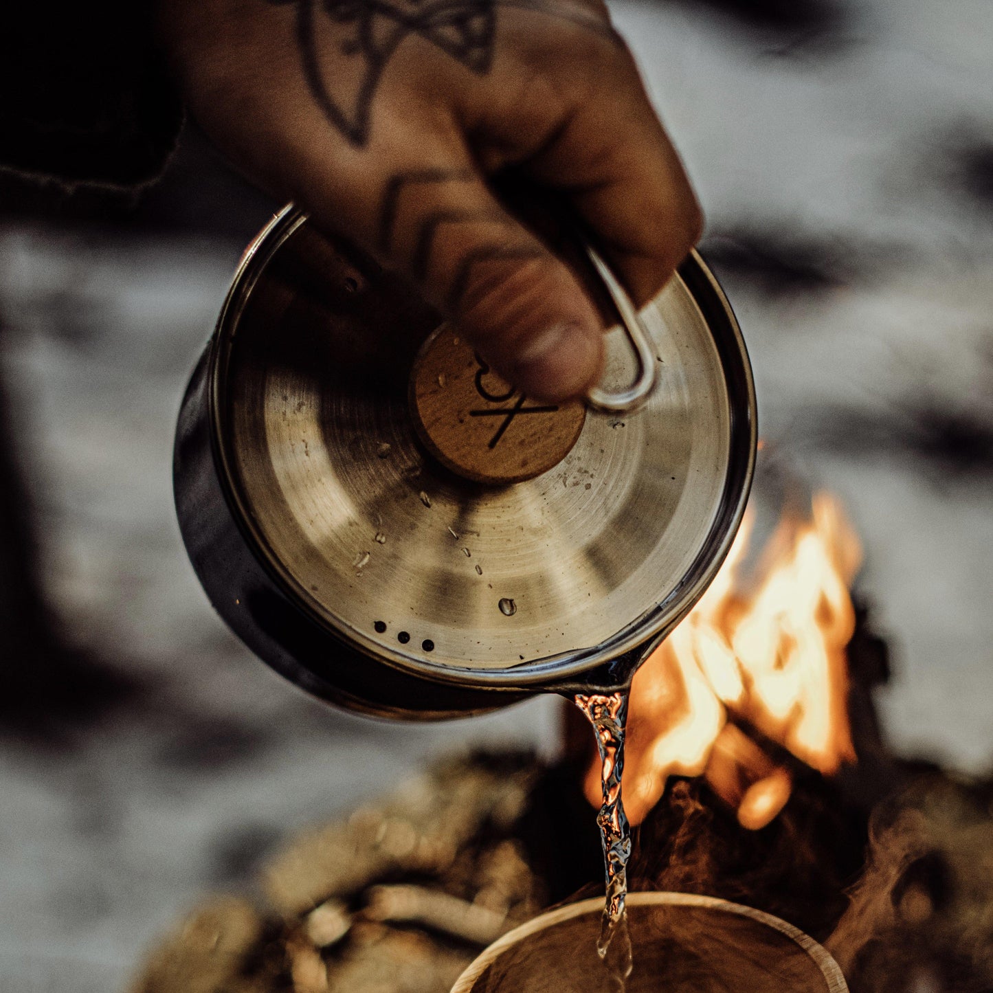 A tattooed hand pours liquid from the Überleben Kessel – The Minimalist Cook Pot into a wooden cup by the campfire—flames flicker, making this ultralight Überleben gear perfect for minimalist backcountry cooking enthusiasts.