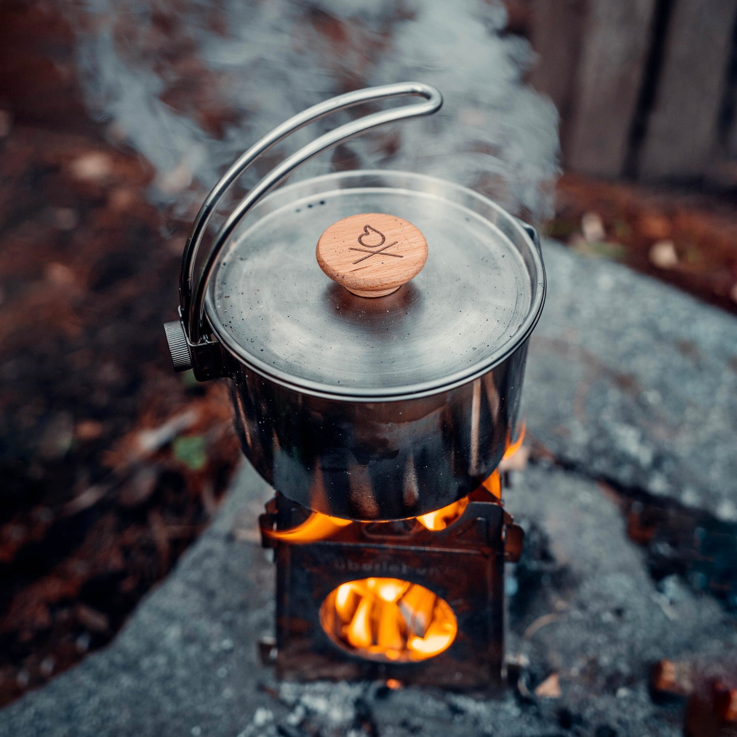 The Überleben Kessel minimalist cook pot by Überleben, featuring a wooden-handled lid, steams while heating over an open flame on a portable stove atop a stone surface outdoors.