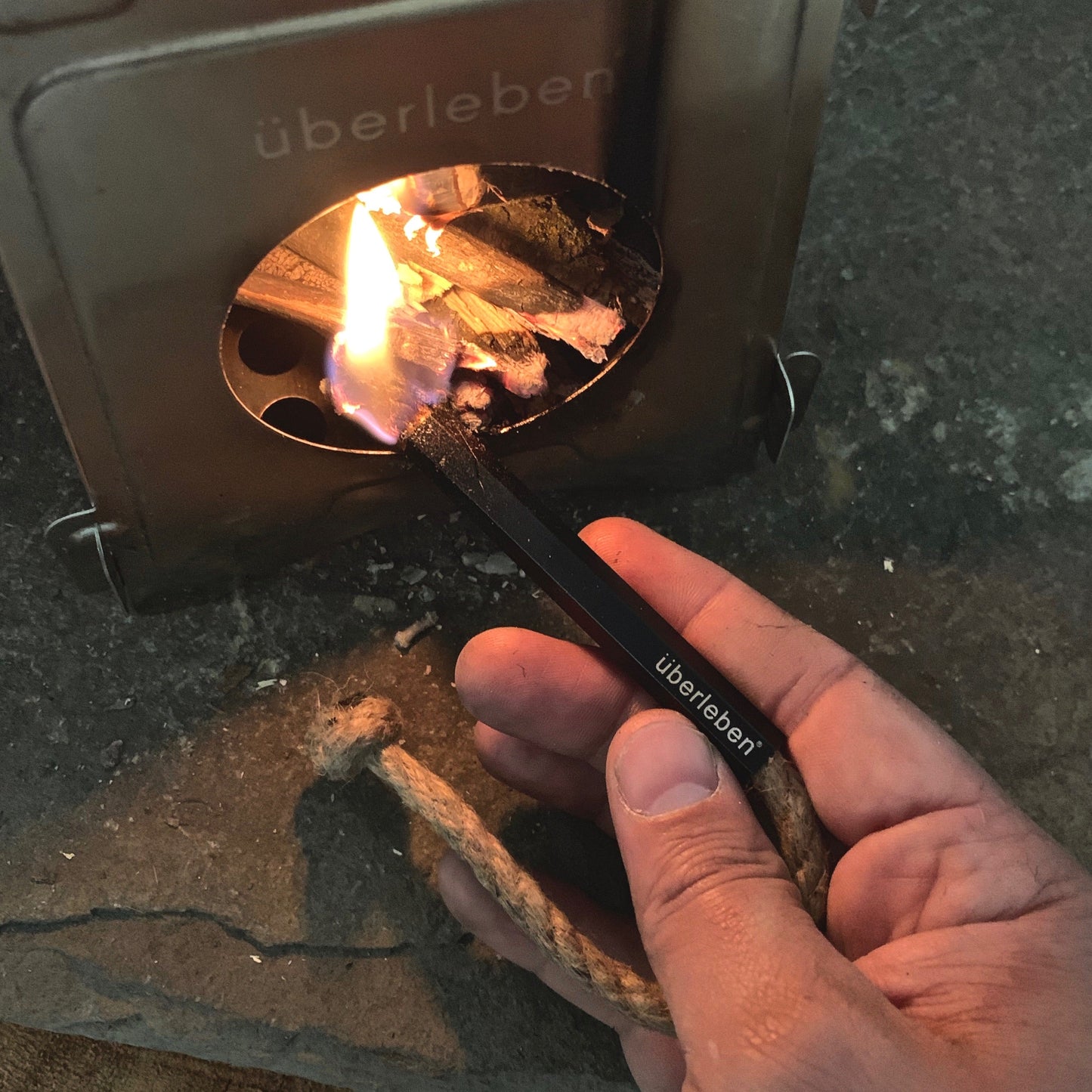 A hand uses the Überleben Tindår | Wick & Bellow Kit to ignite kindling and a waterproof hemp wick inside a small metal stove. As the firestarter is struck, a flame appears and begins catching on the wood pieces in the stove.