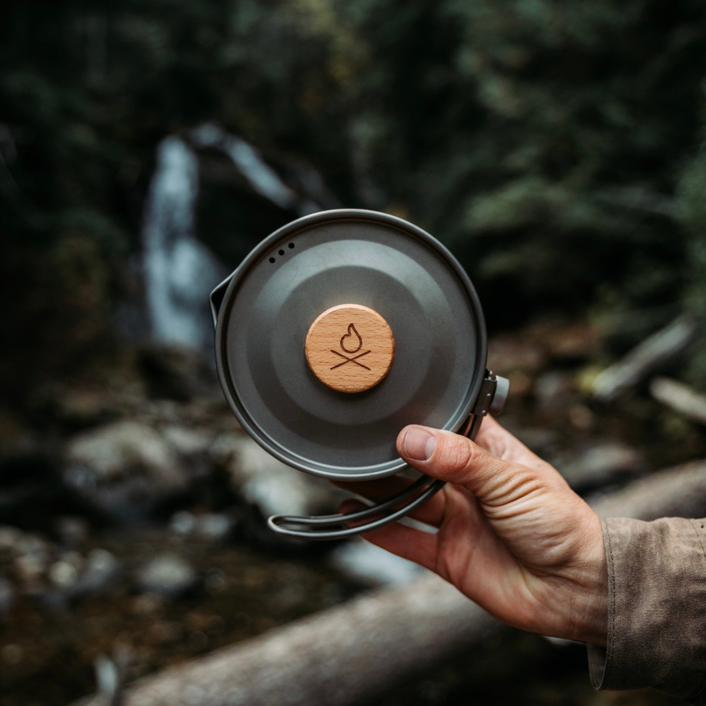 A hand holds the Überleben Kessel – The Minimalist Cook Pot, a round camping mug by Überleben with a wooden lid featuring a campfire symbol, ideal for ultralight backcountry camping, set against a blurred forest and waterfall backdrop.