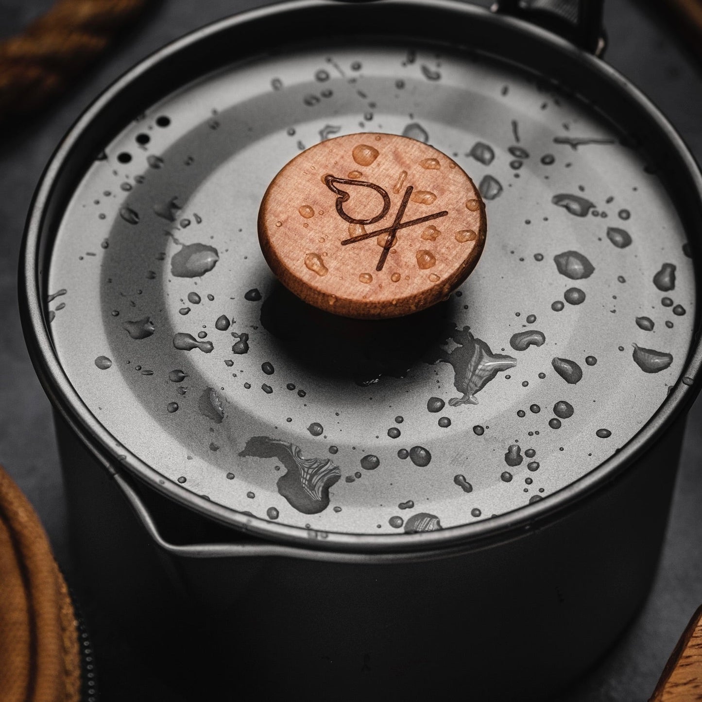 The Überleben Kessel – The Minimalist Cook Pot by Überleben features a metal ultralight design with a decorative engraved wooden lid handle, perfect for backcountry camping cookware needs. Shown with water droplets on a dark surface next to fabric gear.