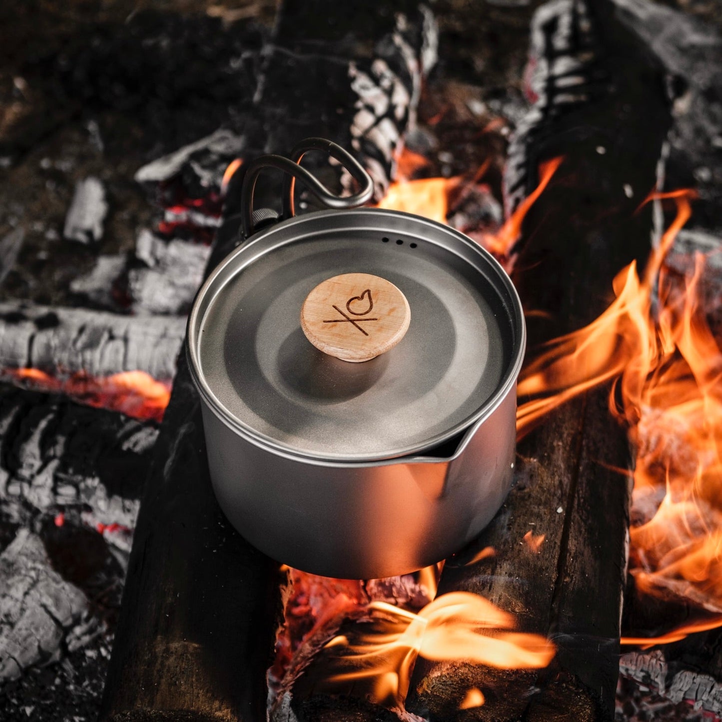 The Überleben Kessel – The Minimalist Cook Pot by Überleben, with a wooden lid and signature heart/X symbol, rests over a campfire with visible flames—ideal minimalist cookware for backcountry cooking or camping adventures.