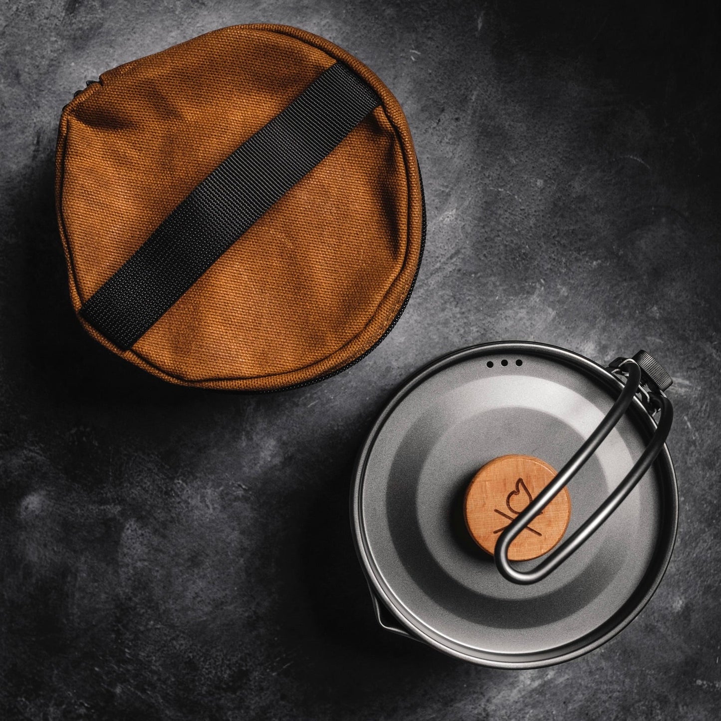 The Überleben Kessel – The Minimalist Cook Pot by Überleben, a gray ultralight pot with a wooden knob and folded handle, is pictured beside a brown fabric bag with black strap on a dark textured surface—ideal for backcountry camping.