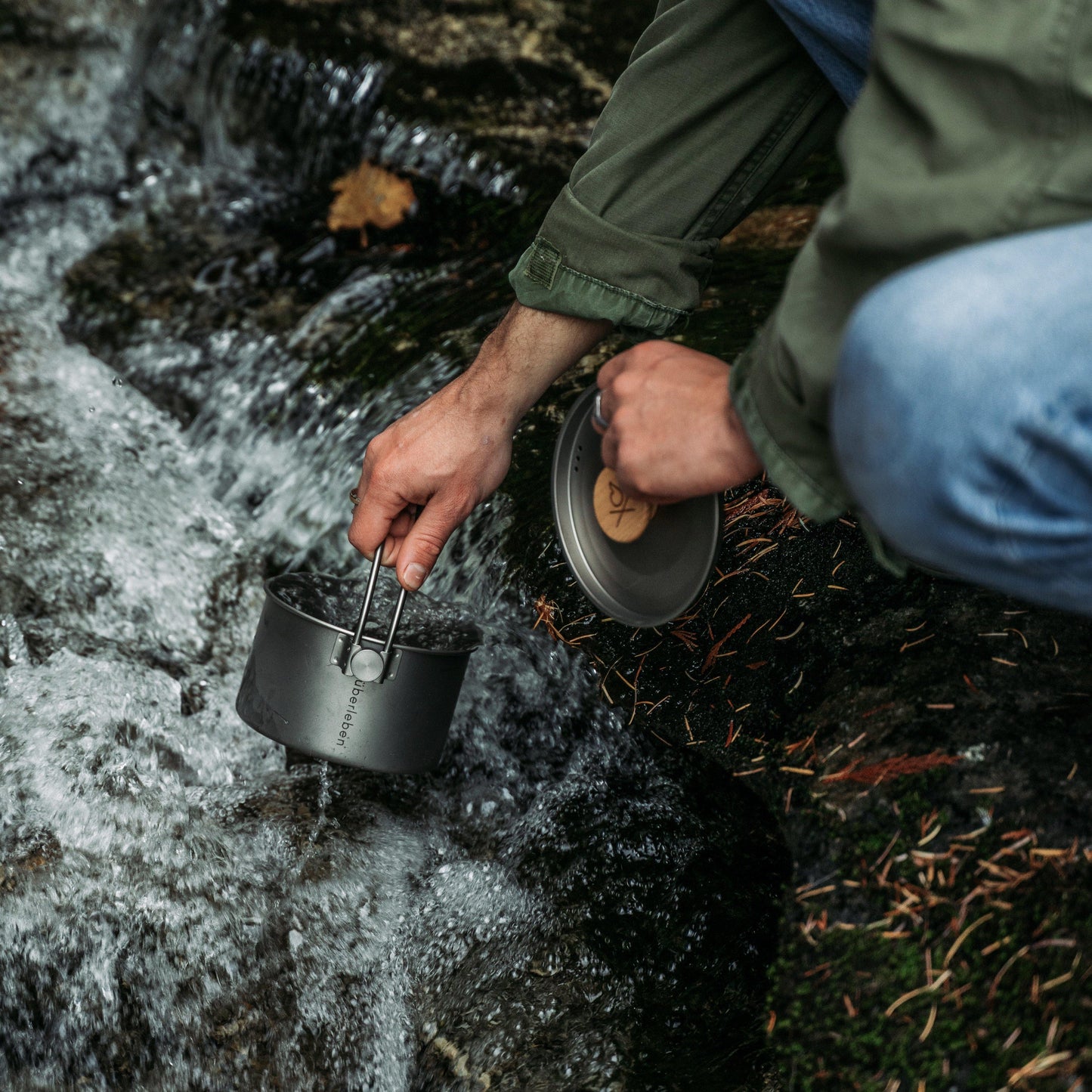 A person in a green jacket collects water from a stream with the Überleben Kessel – The Minimalist Cook Pot by Überleben, holding the pot in one hand and its lid in the other, surrounded by rocks and flowing water.
