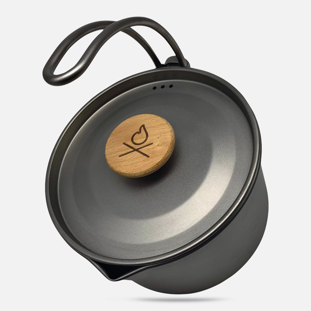The Überleben Kessel – The Minimalist Cook Pot by Überleben is a gray titanium pot with a curved handle and engraved wooden lid knob. It features a snug-fitting lid, small spout for pouring, and is set against a plain white background.
