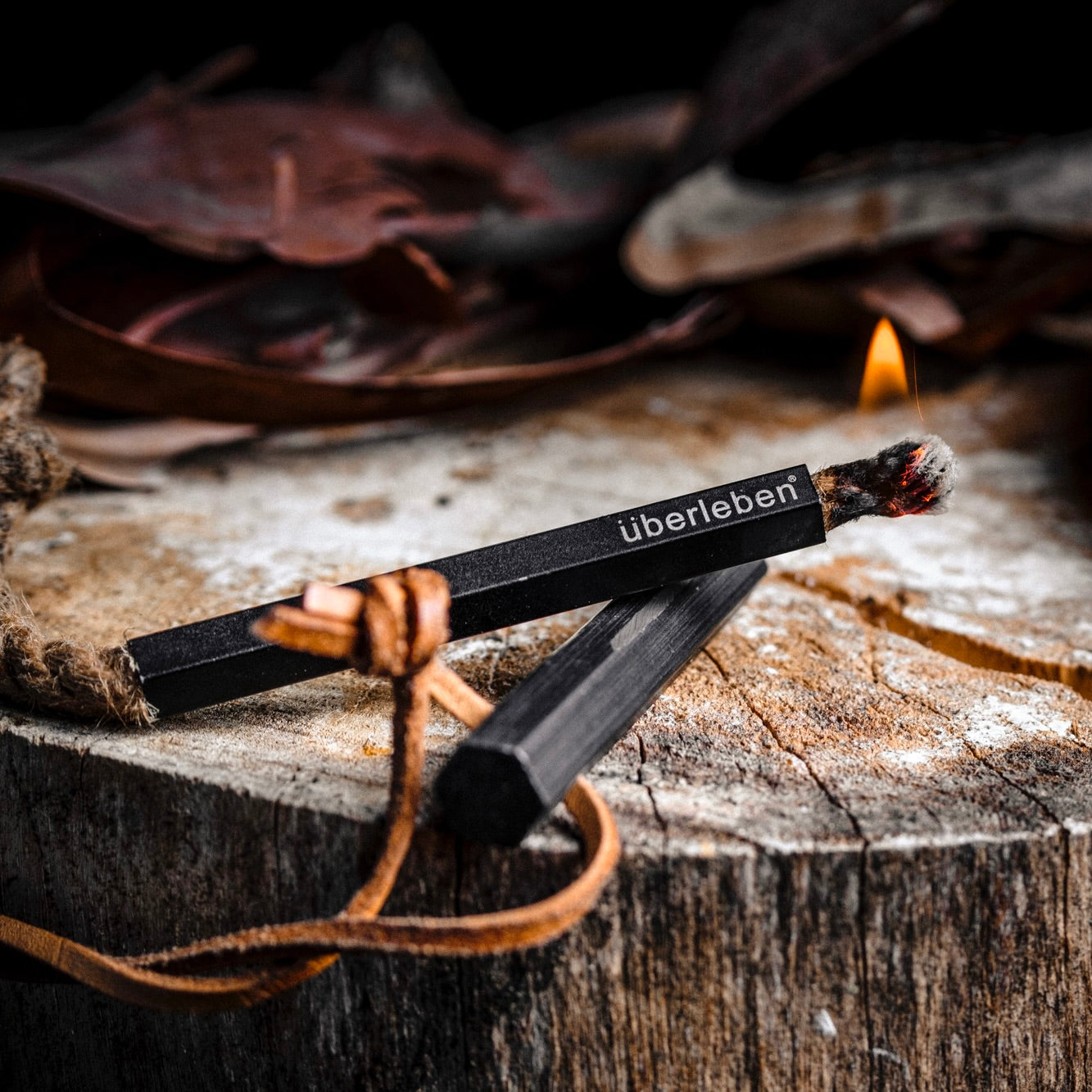 A weatherproof black Überleben Tindår Wick & Bellow Kit and a burning hemp wick rest on a tree stump, surrounded by dried leaves and a leather cord.