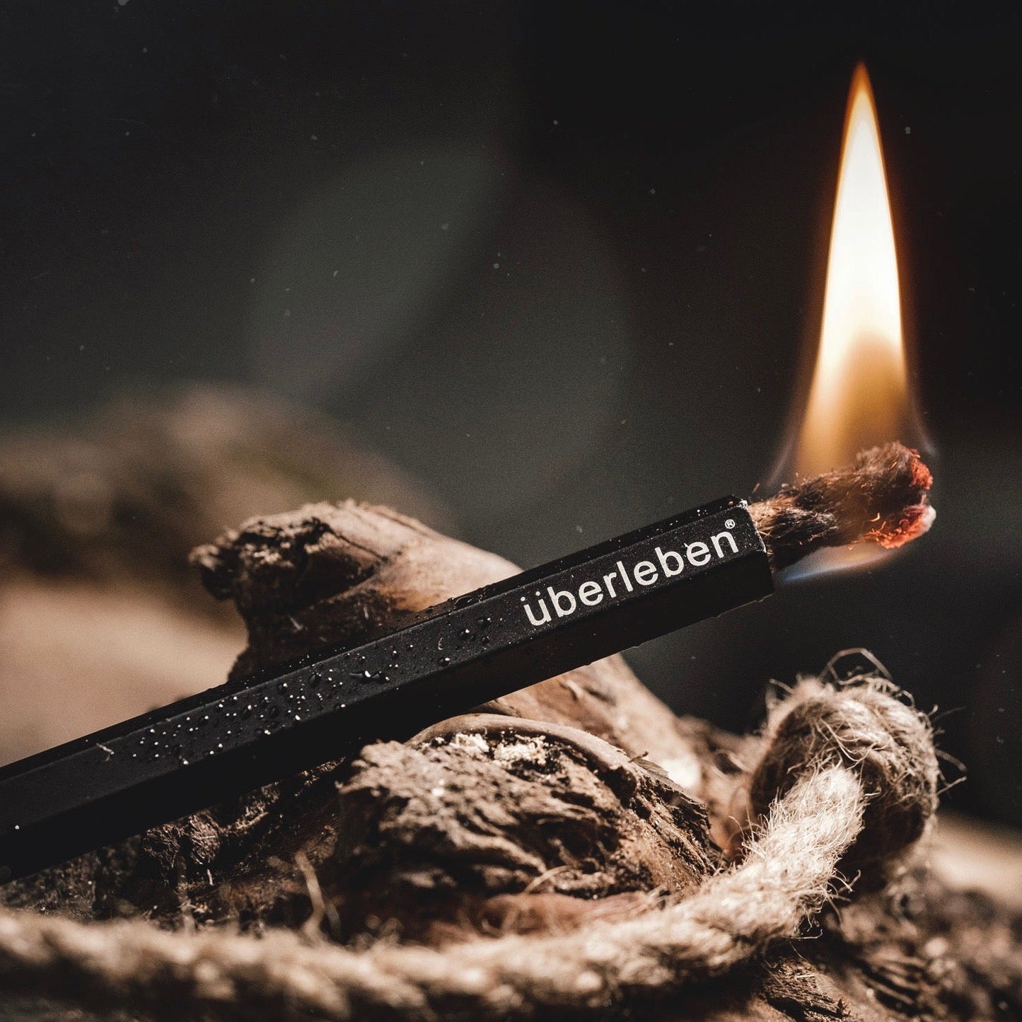 A close-up of the Überleben Tindår | Wick & Bellow Kit igniting its waterproof hemp wick, with a bright flame and wood in the background.