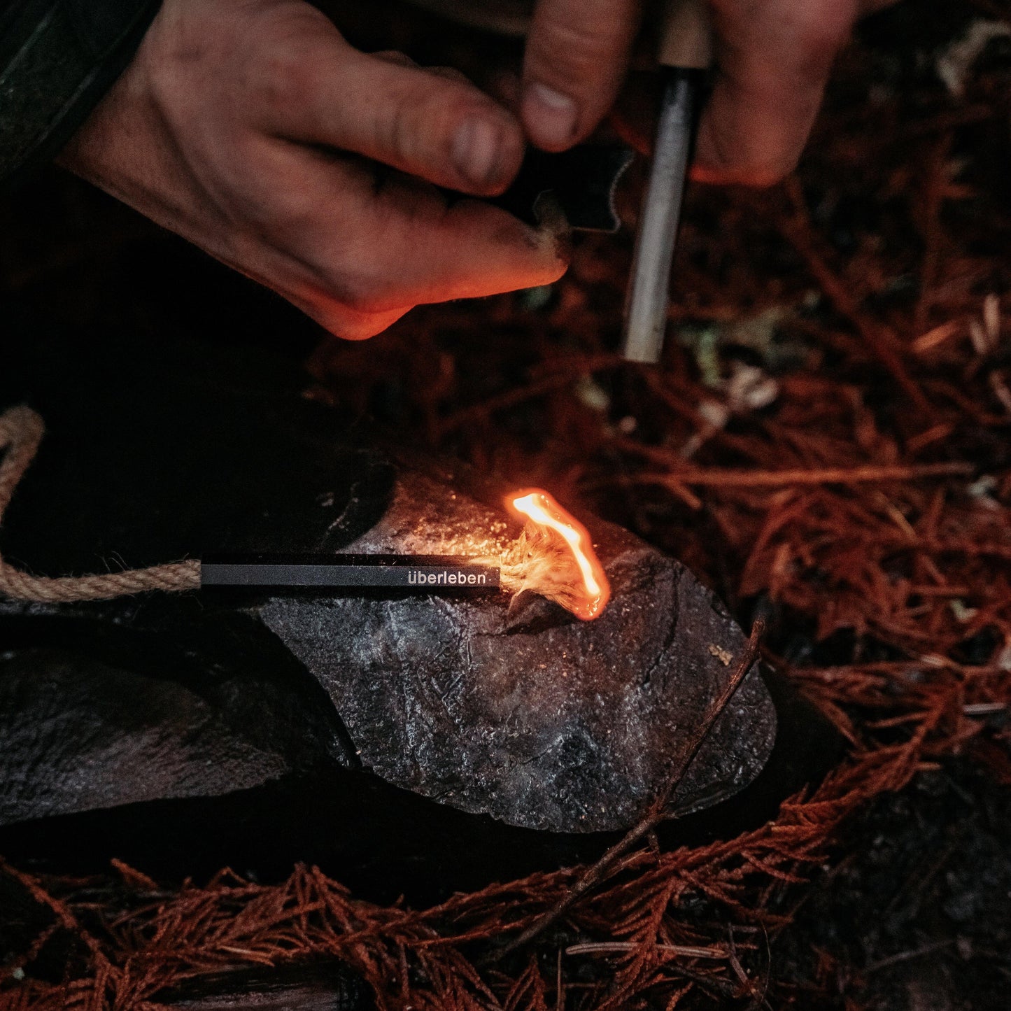 A close-up of hands using the Überleben Tindår Wick & Bellow Kit to ignite a firestarter on a rock, surrounded by brown pine needles.