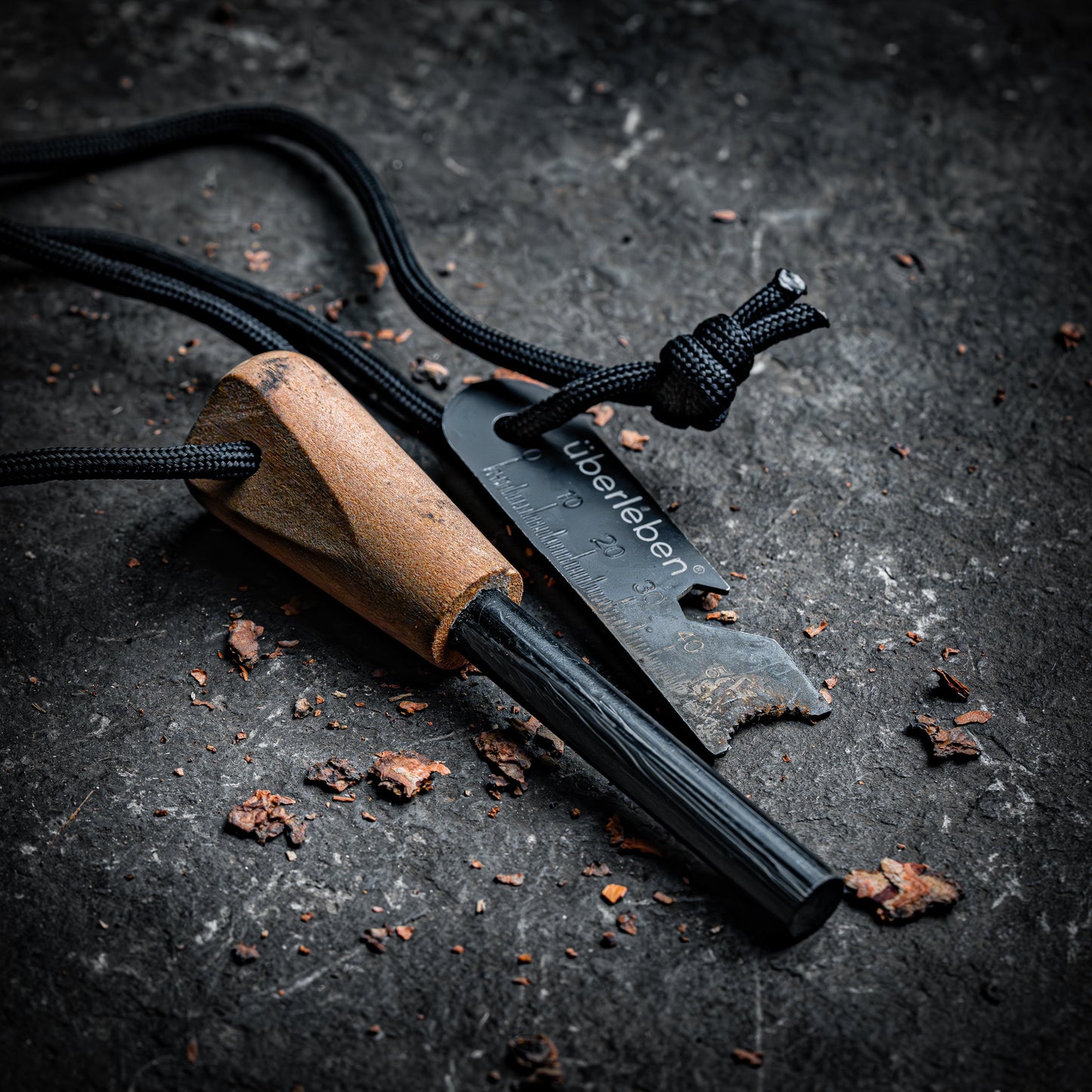 The Überleben Zünden Original – Bushcraft Fire Starter by Überleben features a ferro rod with a wooden handle and black striker, both on a black cord, set on a rugged surface—an essential survival tool for any adventure.