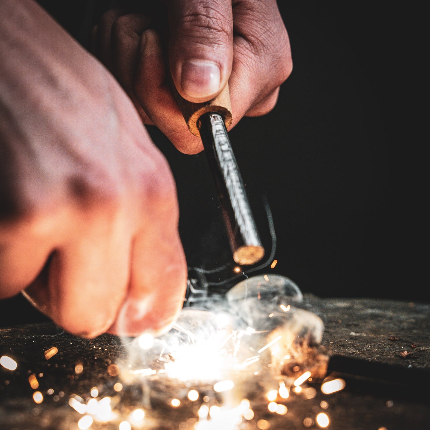 Close-up of hands striking the Überleben Zünden Original Bushcraft Fire Starter by Überleben, creating bright sparks and smoke against a dark background.