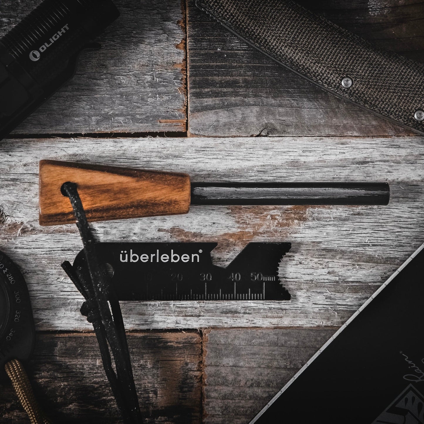 The Überleben Zünden Original – Bushcraft Fire Starter, featuring a wooden handle and black striker with “Überleben” branding, sits on a rustic wooden surface alongside other essential camping survival tools.