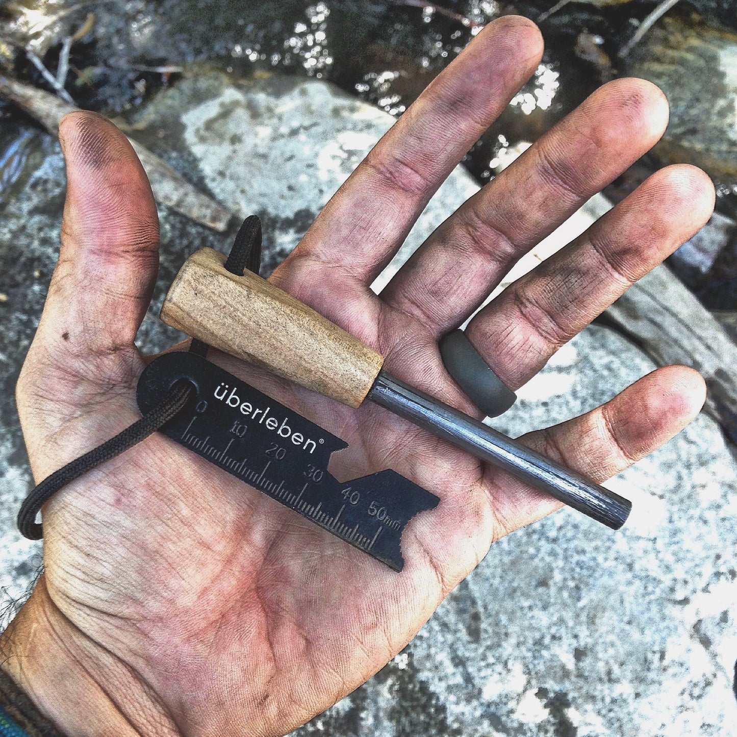 A dirty hand holds the Überleben Zünden Original – Bushcraft Fire Starter by Überleben, featuring a metal ferro rod with wooden handle and black striker, outdoors near rocks and running water—an essential survival tool.