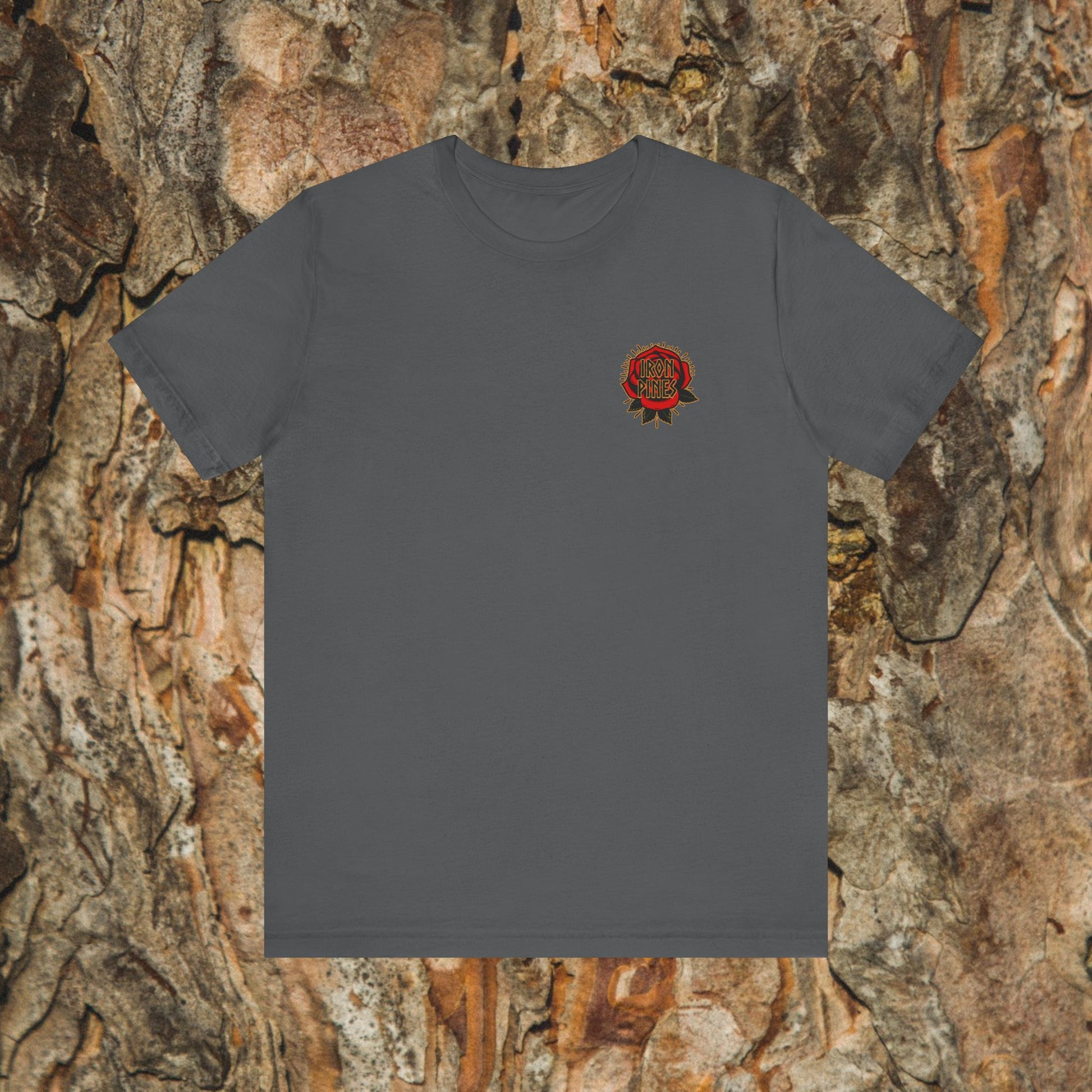 The Never Mistake My Kindness For Weakness Tee by Printify is a unisex charcoal gray graphic t-shirt with a small red rose and text on the left chest, shown against a tree bark background.