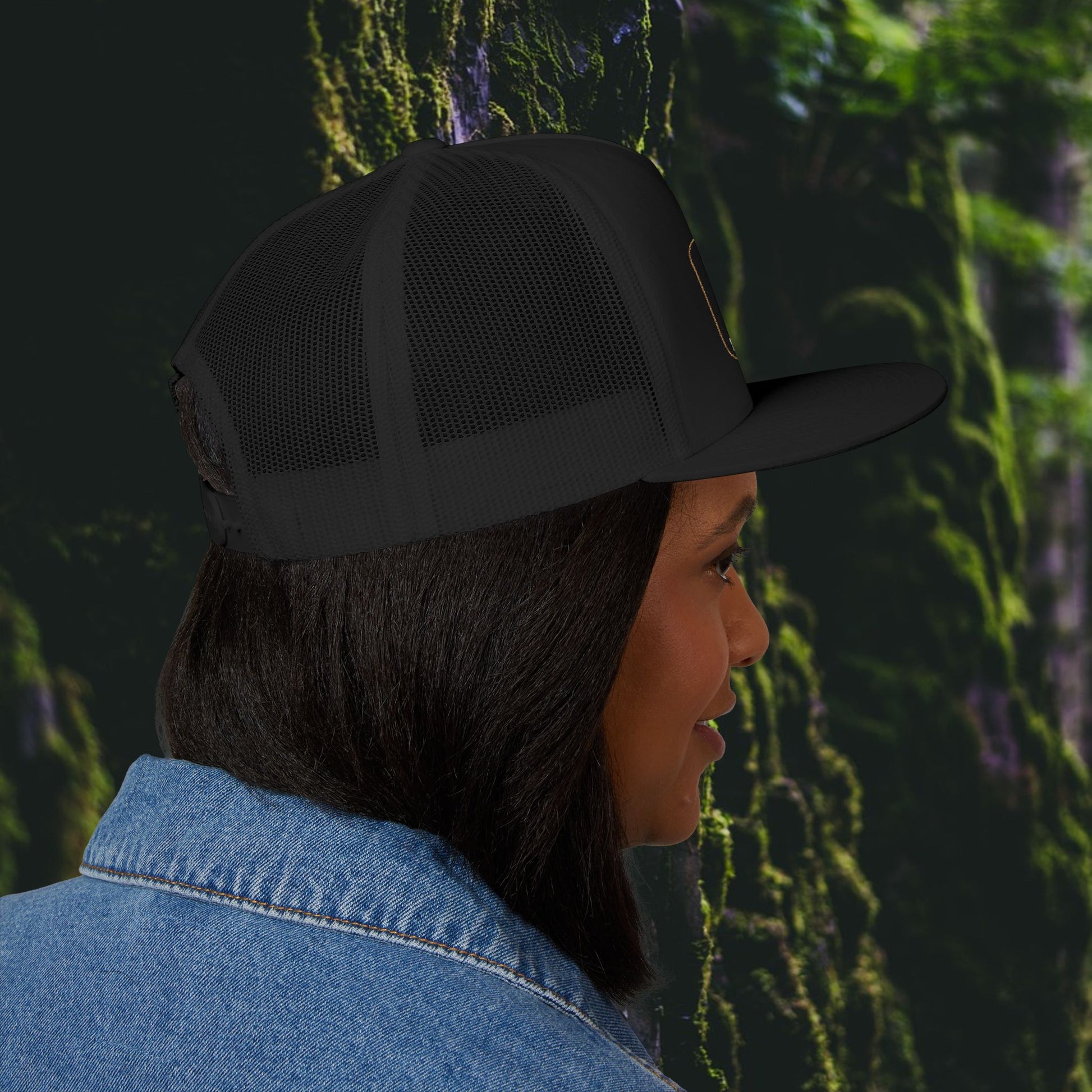 A woman with straight dark hair wears the Printify Iron Pines Trucker Hat – “Live Loud. Be Inconvenient.” Brass Knuckle Edition and a denim jacket, shown in profile against a blurred green, mossy outdoor background.