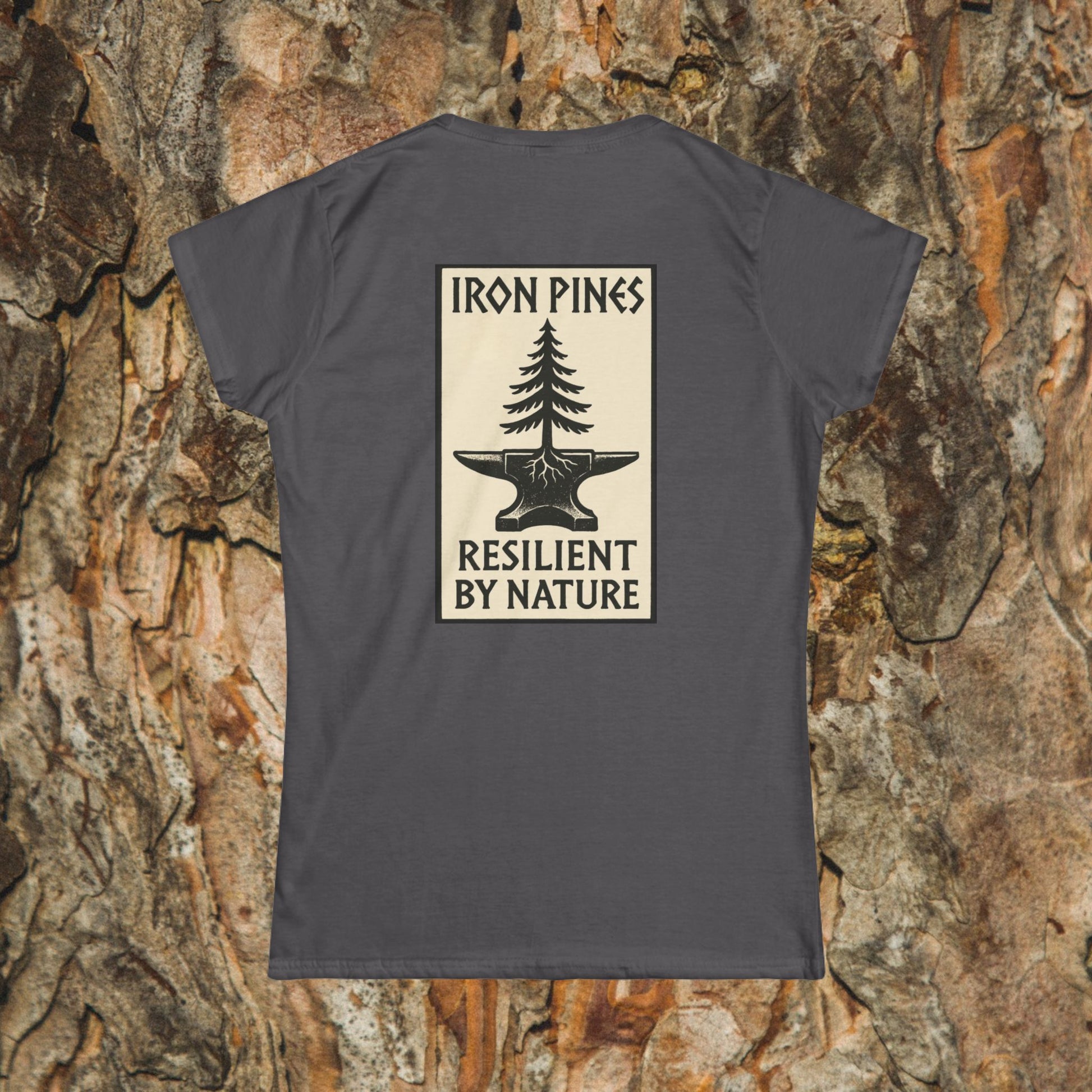 The Iron Pines Womans Tee by Printify features a women’s softstyle fit with a rectangular pine tree on an anvil graphic, “IRON PINES” above and “RESILIENT BY NATURE” below, set against a bark background.