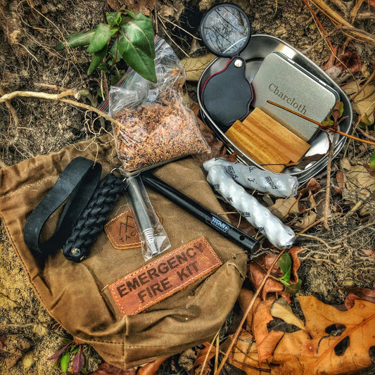 A TITAN Survival TACAMO 15-Piece Emergency Fire Kit with a brown waxed canvas bag sits on the ground, featuring fire starter tools like a ferro rod, paracord, fatwood, charcloth tin, magnifier, and tinder among autumn leaves.