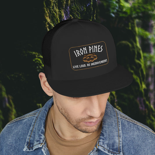 A man in a denim jacket looks down, wearing the Printify Iron Pines Trucker Hat – “Live Loud. Be Inconvenient.” Brass Knuckle Edition, featuring a bold embroidered patch. A blurred forest forms the background.