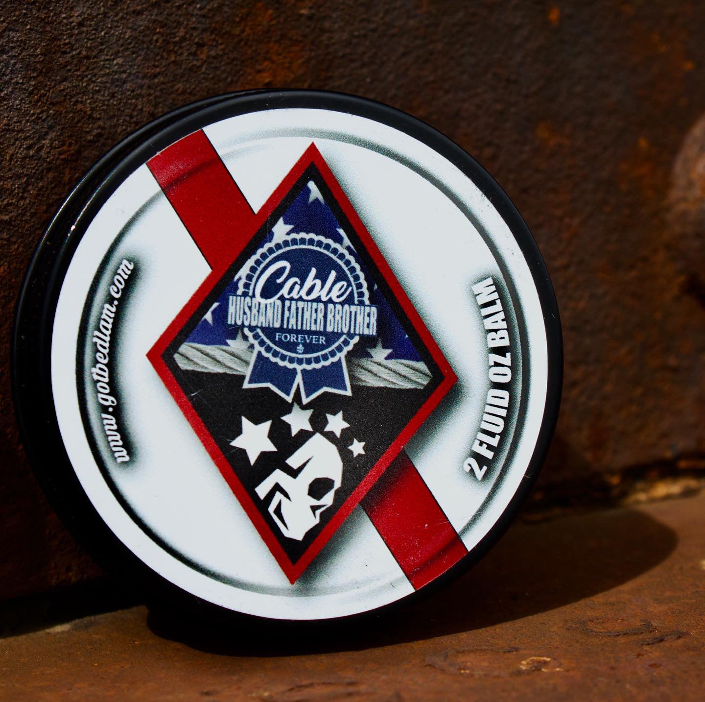 A round tin of CABLE - Warm & Spicy Blend beard balm by Bedlam Beard Company sits on a rusty metal surface, with a white label featuring a red diagonal stripe, blue-black graphics, stars, 2 FLUID OZ BALM, and www.gobeldam.com.