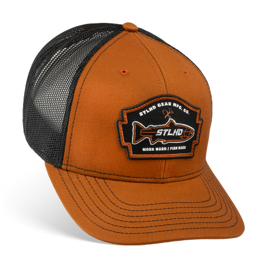 The STLHD GEAR Hook X Hammer Trucker Hat has a brown and black color scheme, breathable mesh back, adjustable snapback, and features a front patch with STLHD GEAR MFG CO. WORK HARD / FISH HARD and a fish graphic with crossed fishing rods.