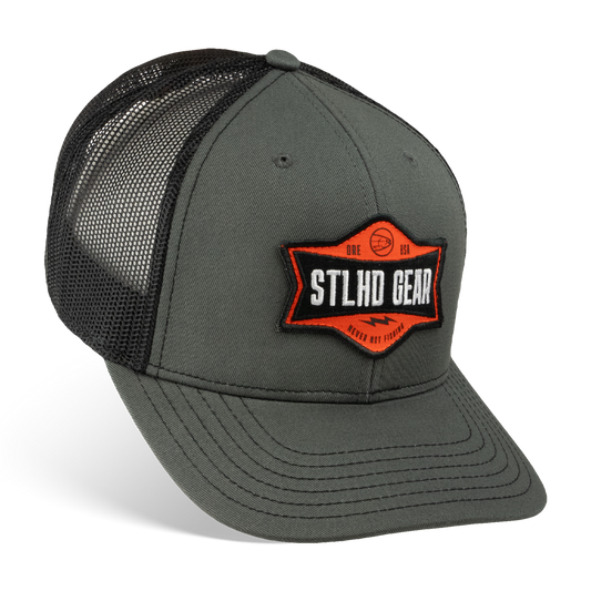 The STLHD GEAR Never Not Fishing Trucker Hat features solid gray front panels, black mesh sides and back, a curved bill, an adjustable snapback closure, and an orange, black, and white STLHD Gear patch on the front.