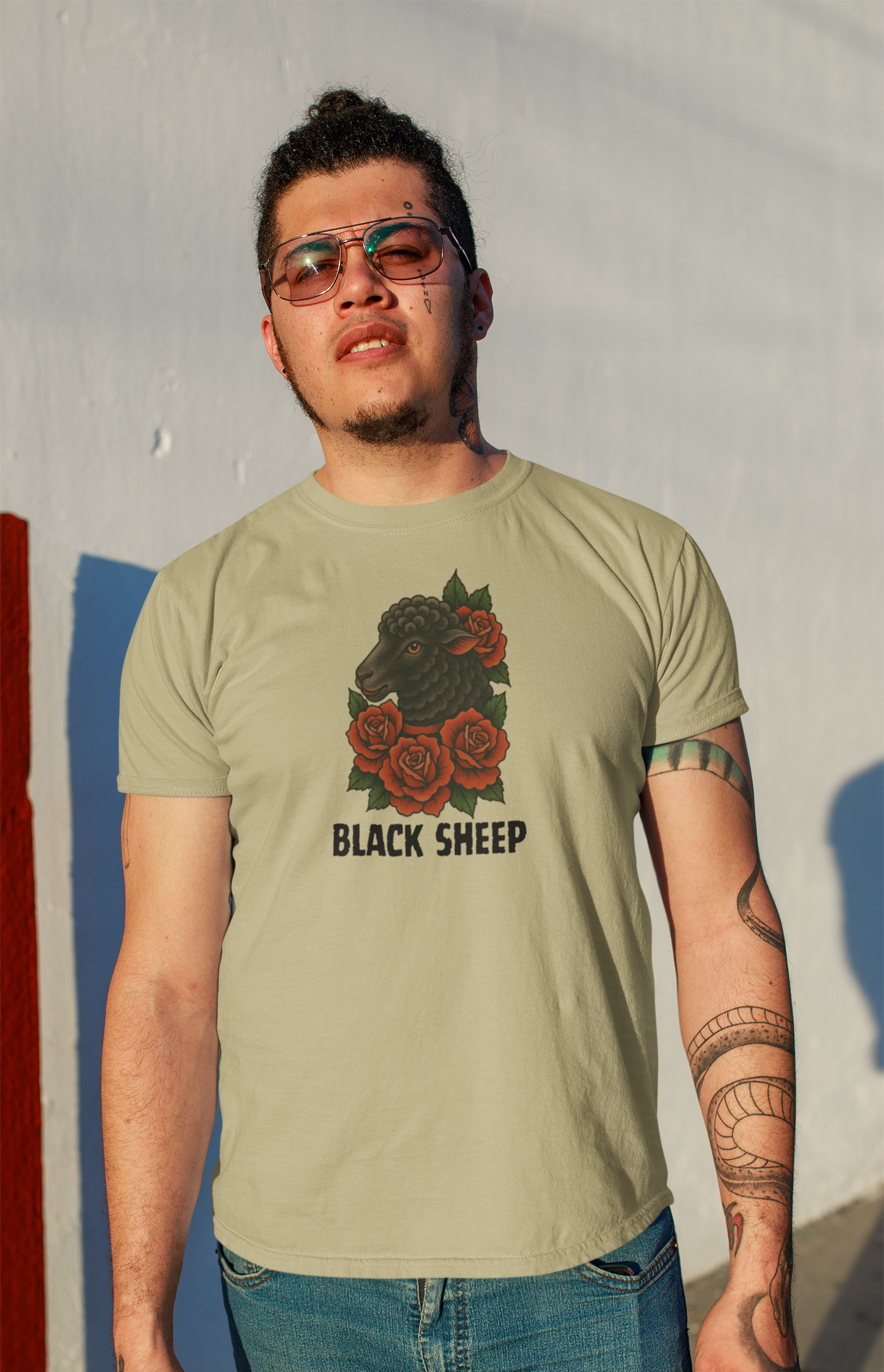 A person with curly hair and glasses stands against a sunlit wall, wearing the Printify Black Sheep Tee featuring red roses and BLACK SHEEP text. Visible arm tattoos add to the unique style of this standout shirt.