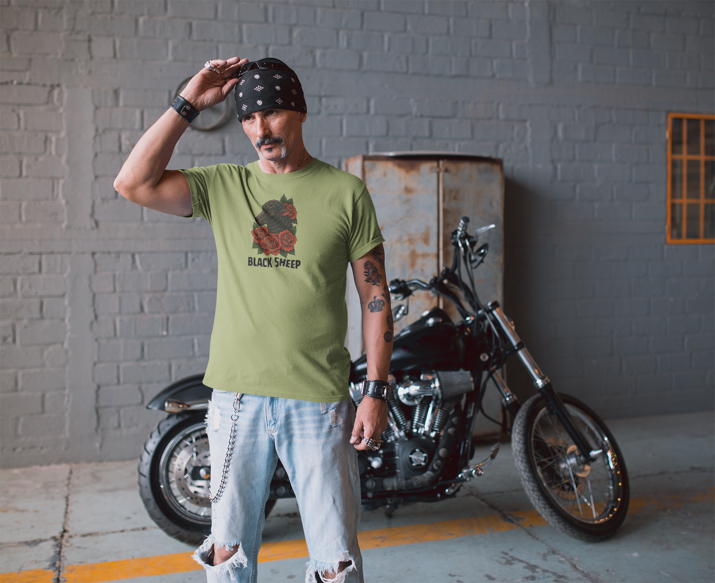 A man in a green Printify Black Sheep Tee, distressed jeans, and a bandana stands by a black motorcycle in an industrial space with gray brick walls and a metal cabinet in the background.