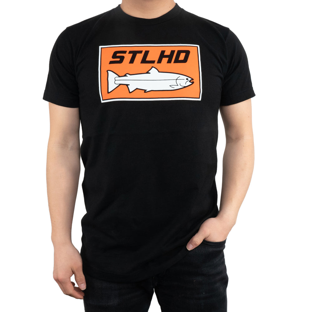 A person wears the STLHD Mens Standard Logo Black T-Shirt by STLHD GEAR, made from CHROMELITE material, with an orange and white rectangle logo showing STLHD above a simple steelhead fish illustration.