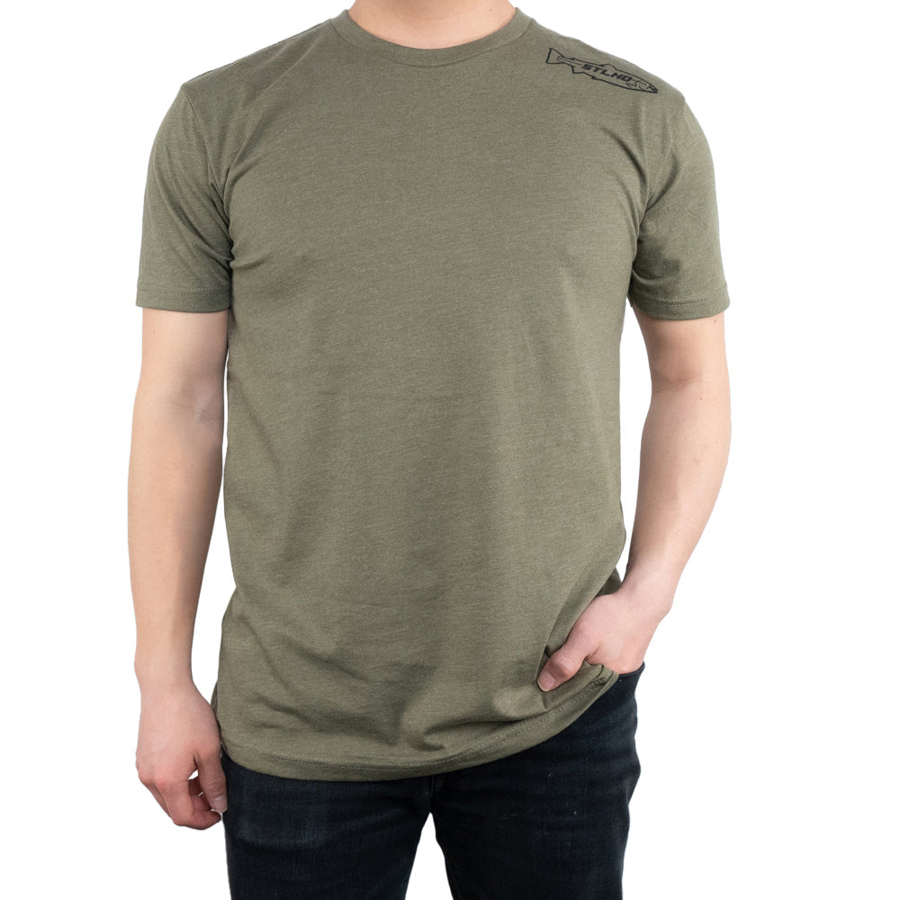 A person wears the STLHD Mens United T-Shirt in Military Green by STLHD GEAR with a small black logo on the left shoulder, paired with dark jeans, right hand in pocket, against a white background.