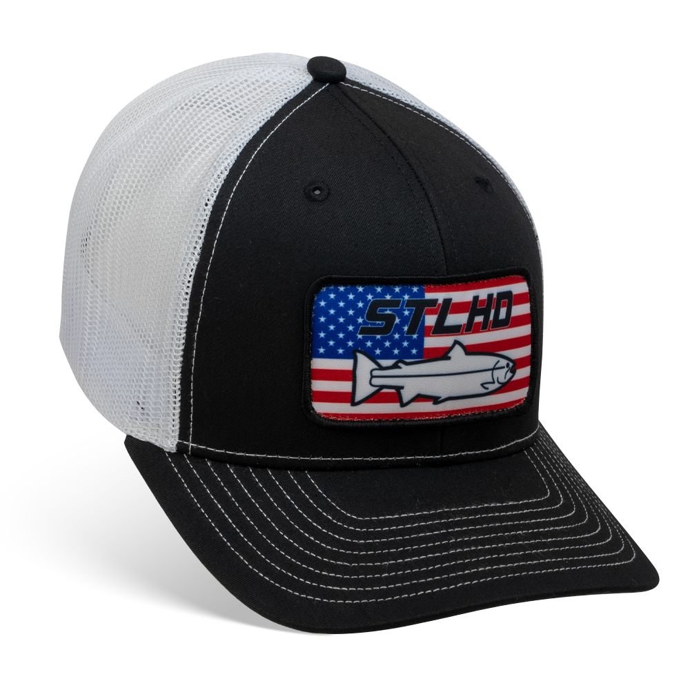 The STLHD GEAR Nation Black/White Trucker Snapback Hat features mesh back, adjustable snapback, an American flag patch, STLHD, and a wild steelhead fish outline on the front.