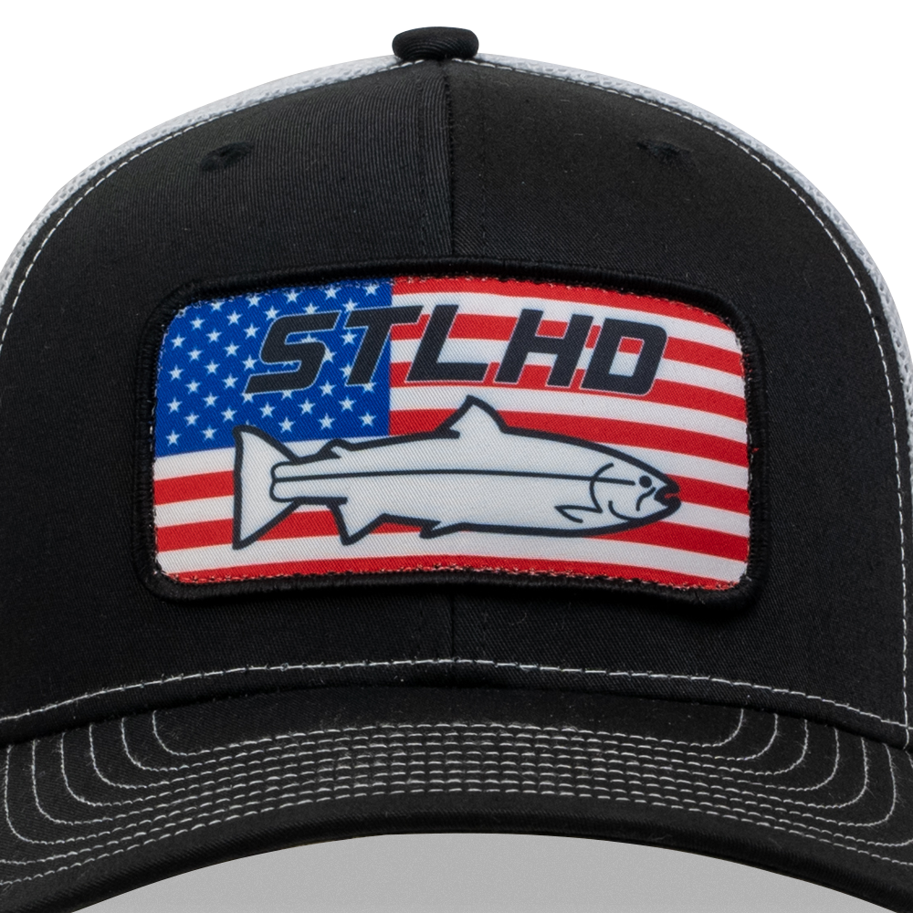 The STLHD GEAR Nation Black/White Trucker Snapback Hat features a patch with the American flag, a wild steelhead outline, and bold STLHD text—an essential accessory for any STLHD fan.
