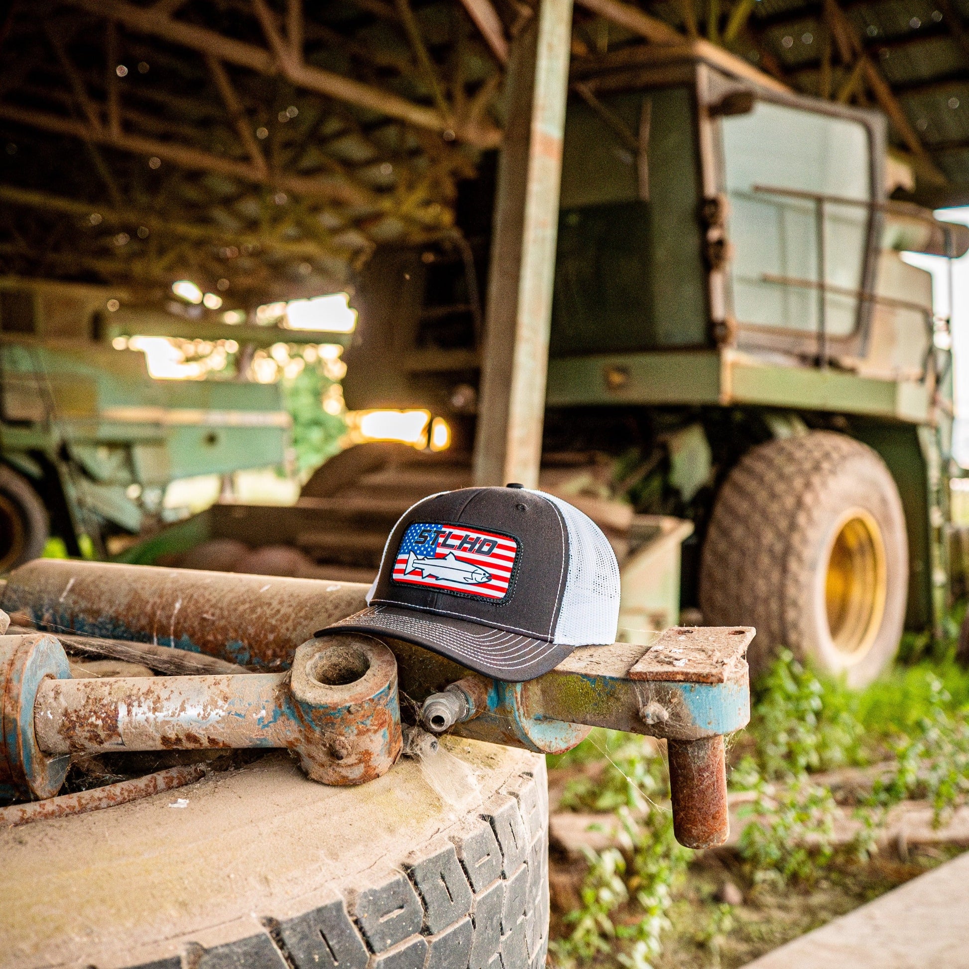 The STLHD GEAR STLHD Nation Black/White Trucker Snapback Hat sits on a large tire in front of an old green combine harvester inside a rustic, open-sided barn.