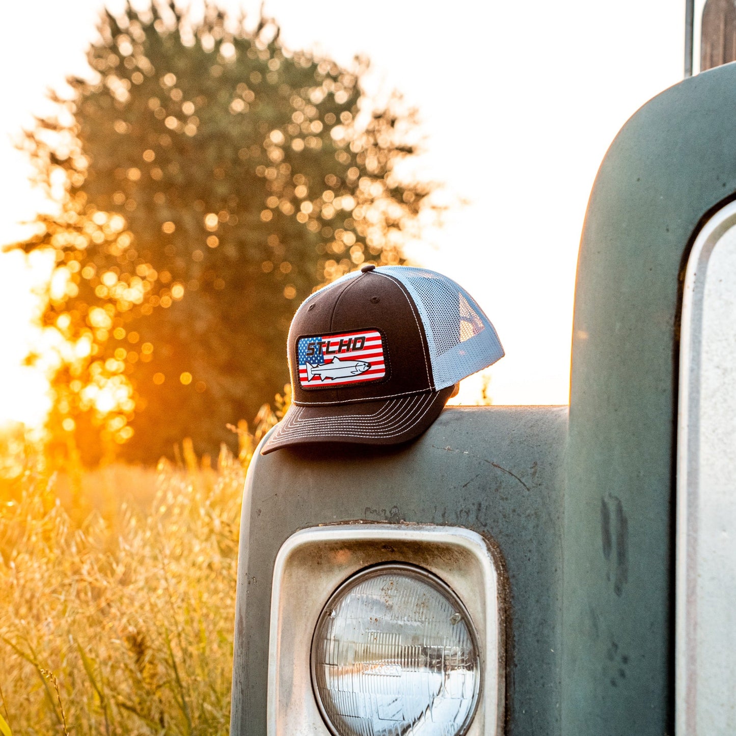 The STLHD GEAR STLHD Nation Black/White Trucker Snapback Hat, featuring an American flag and Chevy truck logo, rests on a vintage vehicle’s headlight in a field at sunset—a proud salute to STLHD Nation.