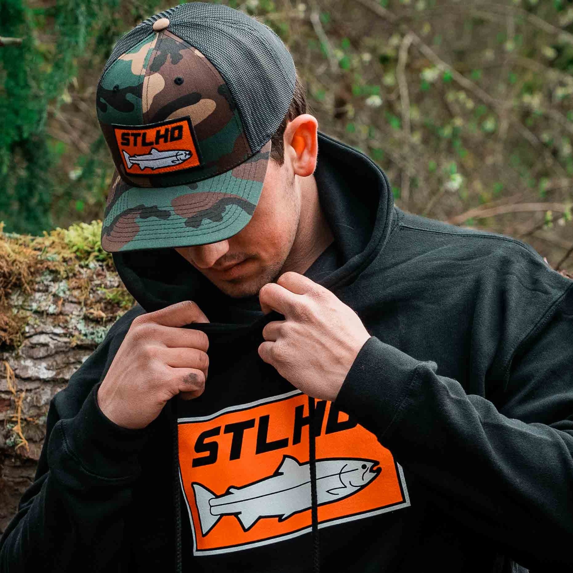 Outdoors, someone wears the STLHD GEAR Camo Snapback Trucker Hat and a black hoodie, both with an orange STLHD patch and fish graphic. In the background, there’s greenery and a fallen tree.
